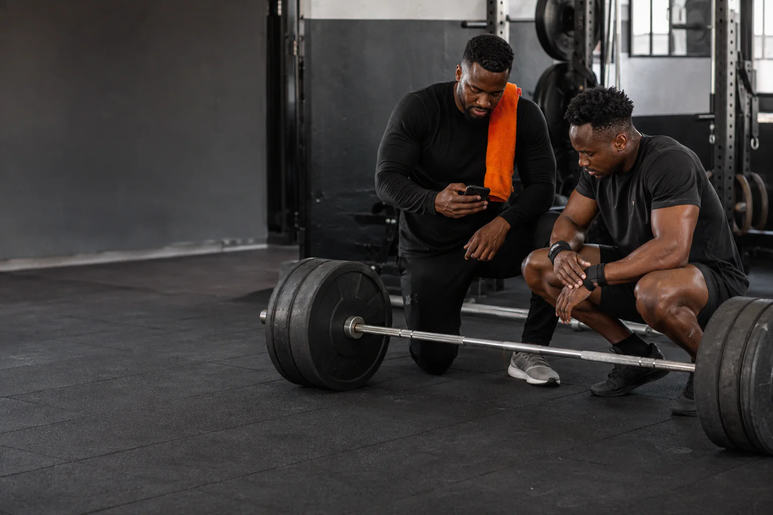 A coach and an athlete checking a session on a phone beside a loaded barbell